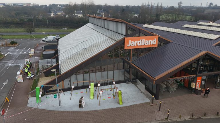 Drone en intervention pour le nettoyage de la grande toiture d’un magasin Jardiland, avec l’équipe au sol sécurisant la zone et préparant les produits, vue aérienne montrant la façade vitrée et le parking du site.